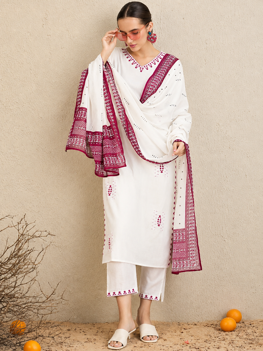 A woman models a white traditional South Asian outfit with magenta embroidered borders and a matching dupatta, standing against a textured beige wall with scattered oranges on the ground.