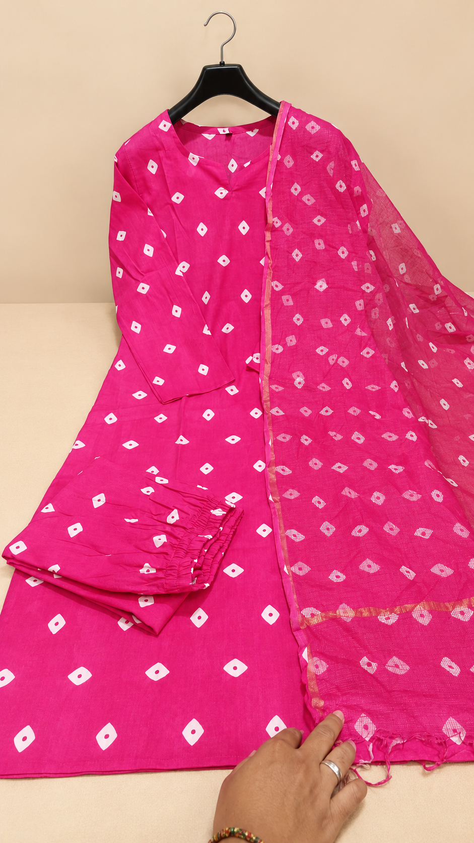 Model wearing a magenta Bandhani print Anarkali kurta with white motifs, matching pants and a sheer bordered dupatta against a beige background.

