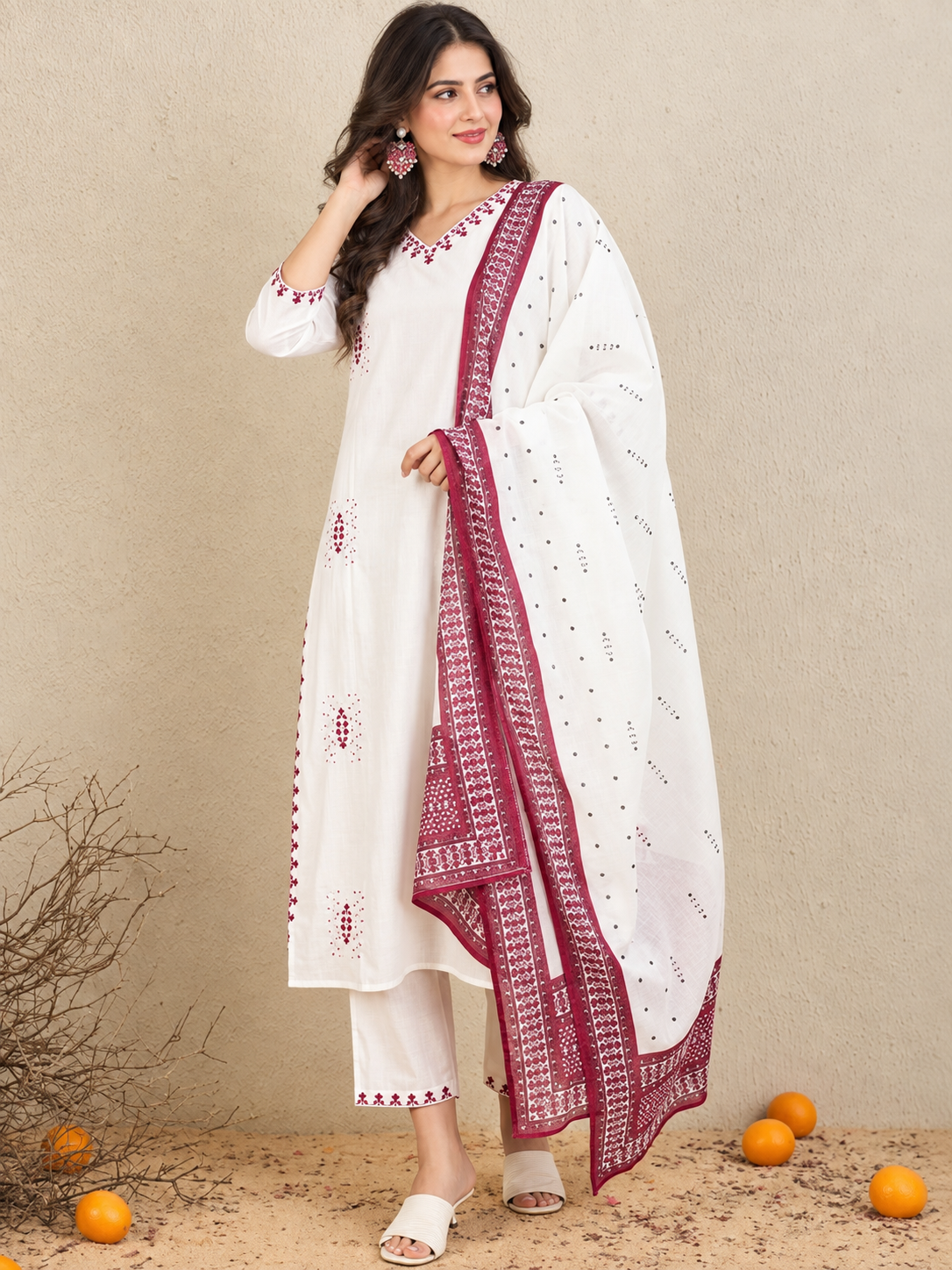 A woman models a white ethnic suit set featuring a kurta with red embroidery, matching trousers, and a white dupatta with a red border, set against a textured beige wall with scattered oranges on the ground.