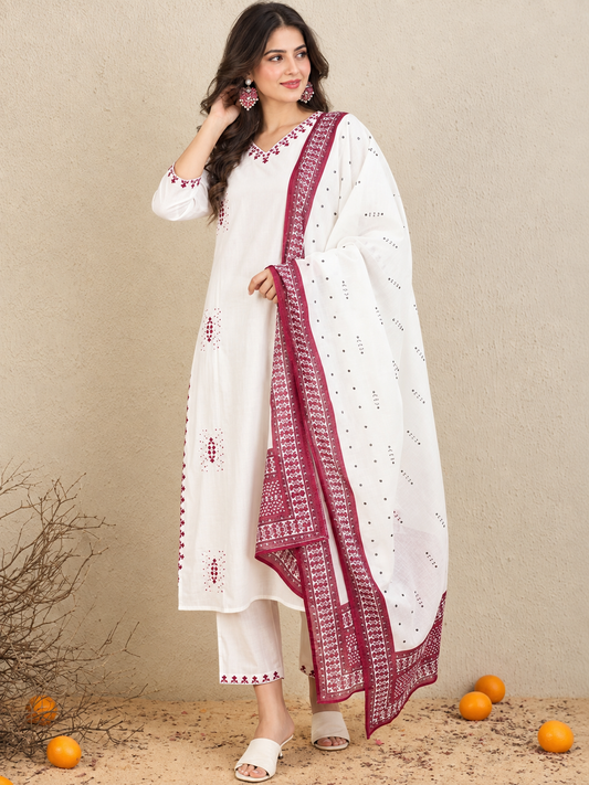 A woman models a white ethnic suit set featuring a kurta with red embroidery, matching trousers, and a white dupatta with a red border, set against a textured beige wall with scattered oranges on the ground.