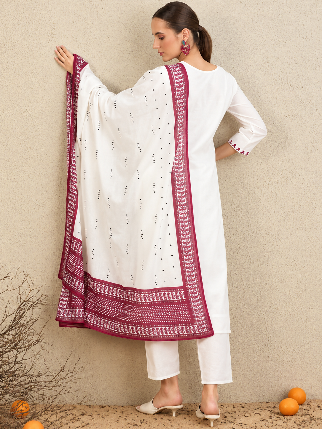 A woman models a white three-piece traditional outfit with a deep magenta border dupatta, standing against a textured beige wall with decorative dried branches and oranges on the floor.