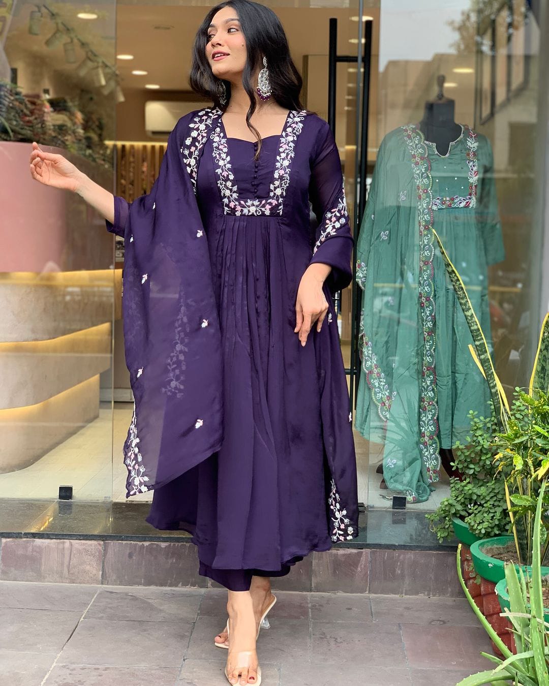 Woman in a purple dress with white embroidery standing in front of a store window displaying green dresses.