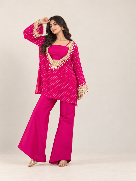 A woman modeling a bright pink traditional outfit with gold embroidered details, consisting of a kurta, matching pants, and a solid pink inner top, set against a plain light beige studio background.