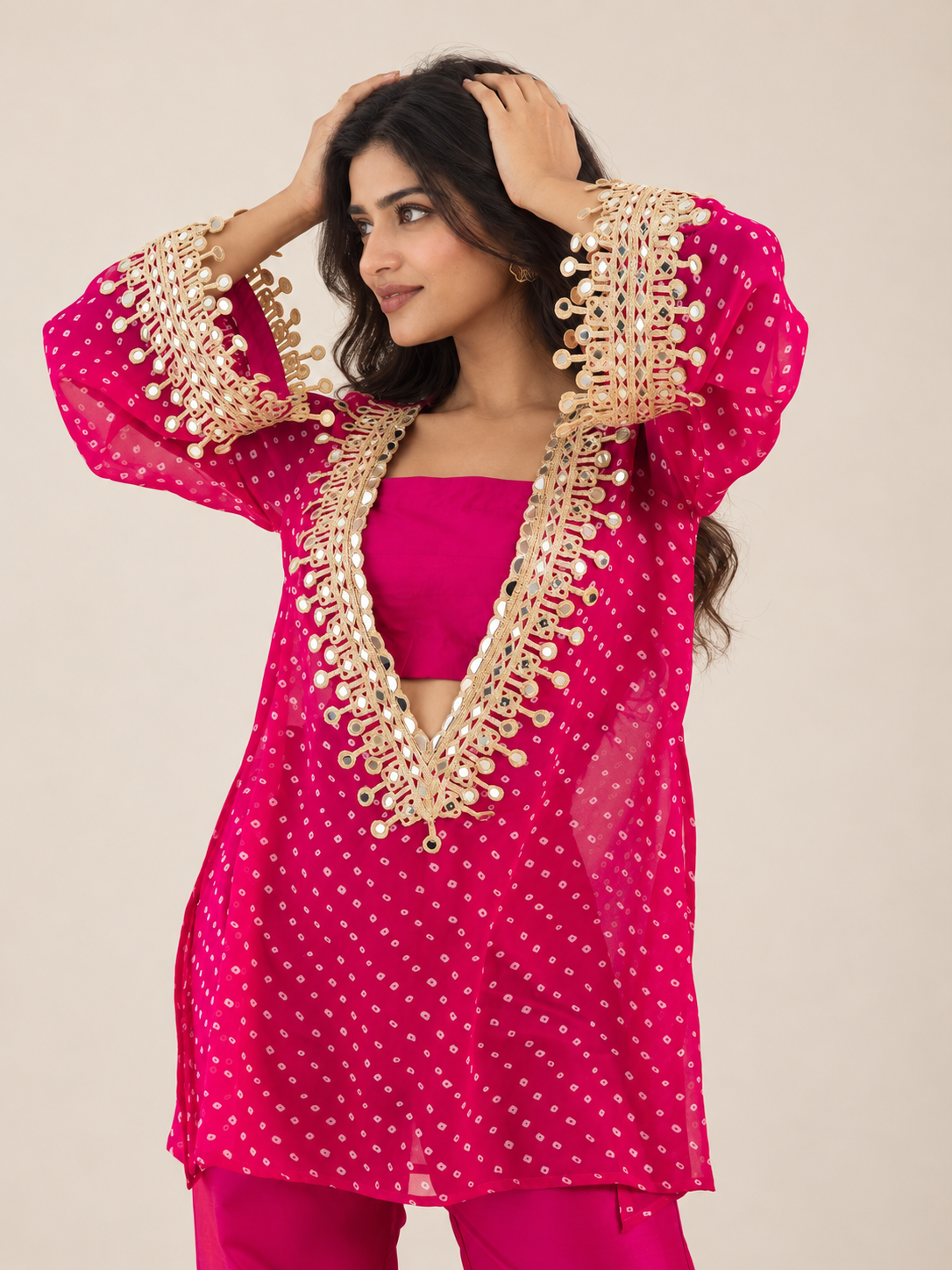 A woman modeling a bright pink traditional outfit with gold embroidery and mirror work on the sleeves and neckline, paired with matching pink pants, against a plain beige studio background.