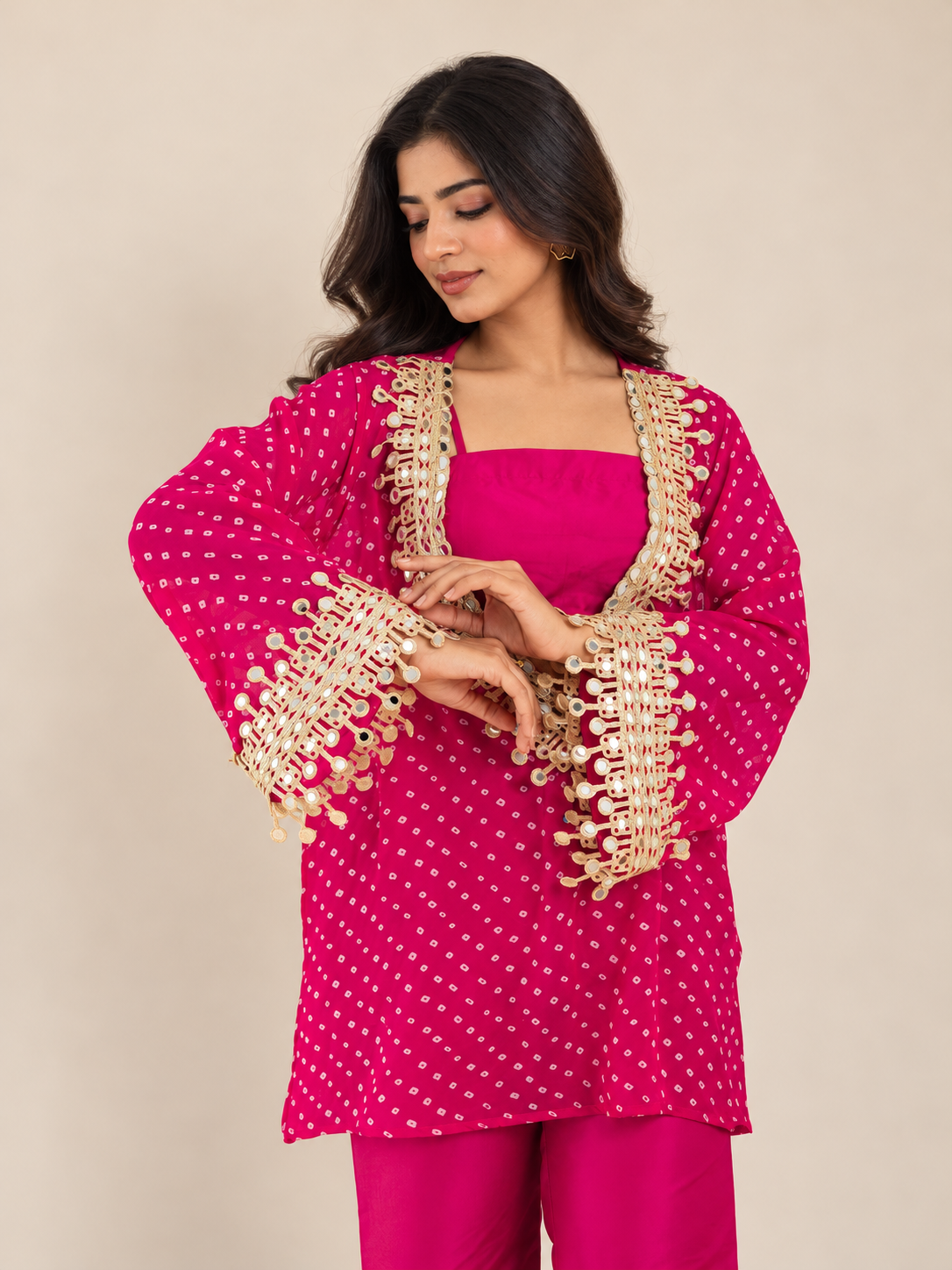 A woman modeling a bright pink traditional outfit with gold coin embroidery on the sleeves and neckline, paired with matching pink pants, against a plain beige studio background.