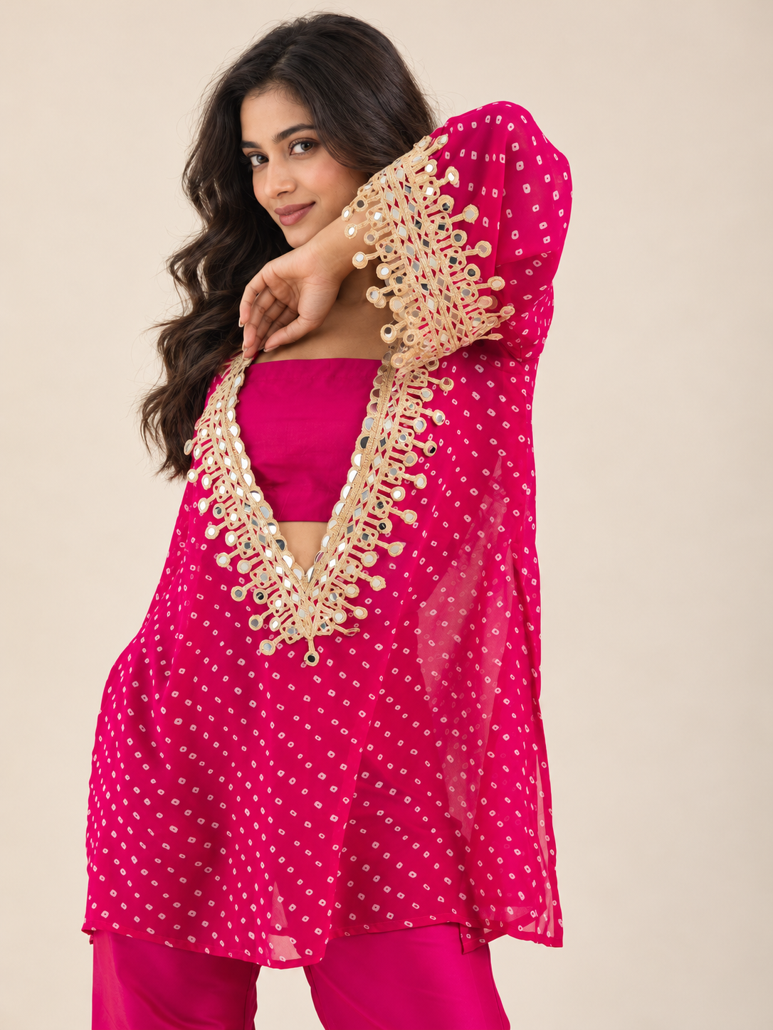 A woman modeling a bright pink traditional outfit with gold embroidered detailing on the neckline, paired with matching pink pants, against a plain beige studio background.