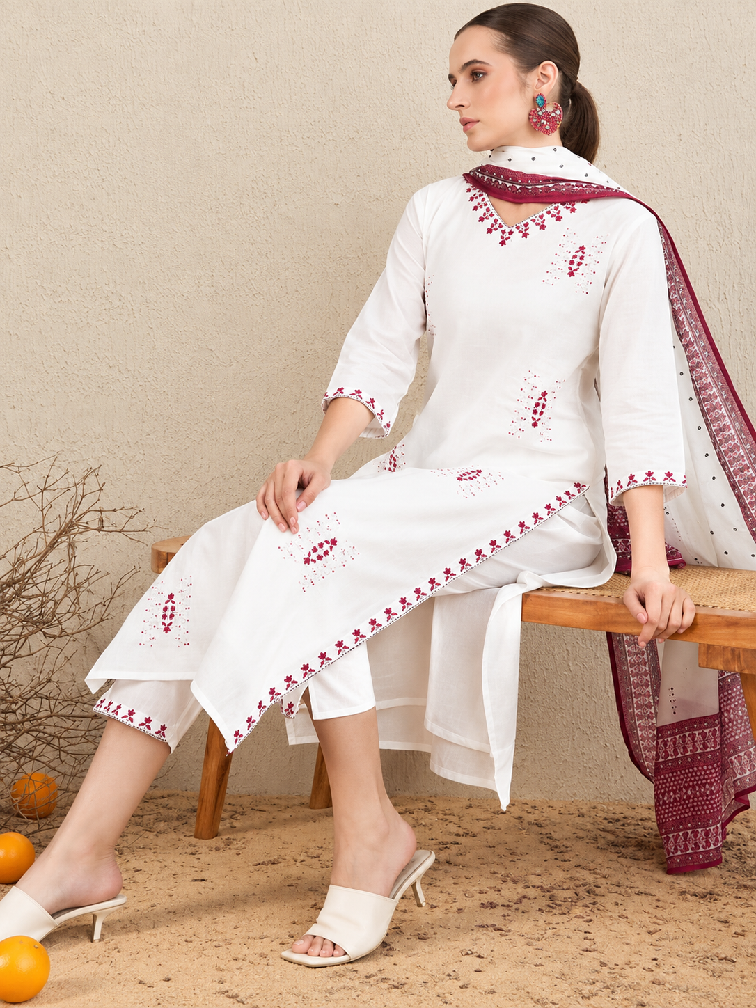 A woman models a white three-piece salwar kameez suit with red embroidered details, paired with a matching red and white patterned dupatta, seated on a wooden bench against a textured beige wall.