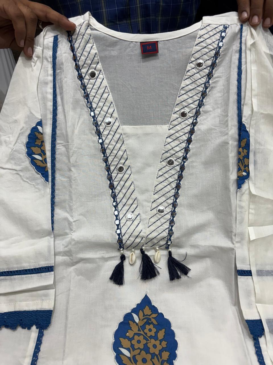 A white cotton kurta with blue and gold embroidery, featuring a deep V-neckline with silver sequins, tassel details, and blue decorative borders. Hands are holding the garment open at the shoulders.