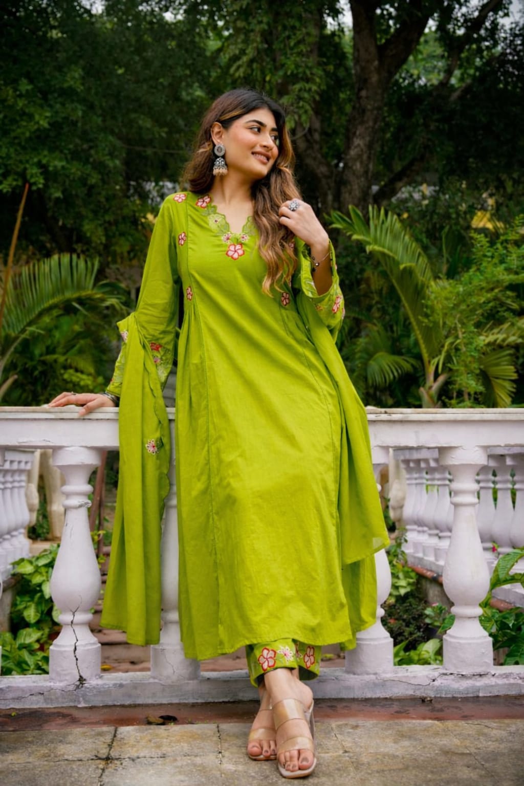 A woman wearing a bright lime green traditional kurta set with floral embroidery, standing on a white stone balcony with lush green garden foliage in the background.