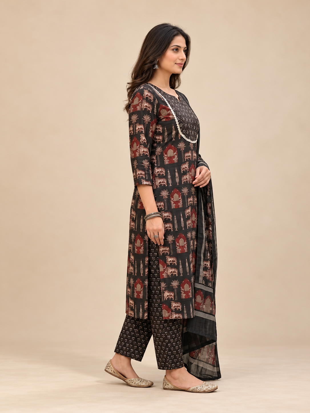 A woman modeling a traditional Indian salwar kameez set featuring a black base with red and gold motifs, paired with matching patterned trousers and a black dupatta, set against a solid beige studio background.