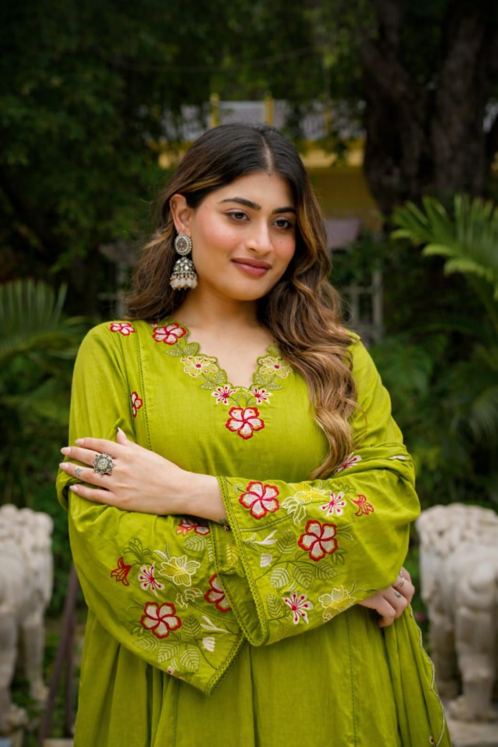 A woman wearing a bright lime green traditional embroidered dress with red and pink floral patterns, standing outdoors in a garden setting.