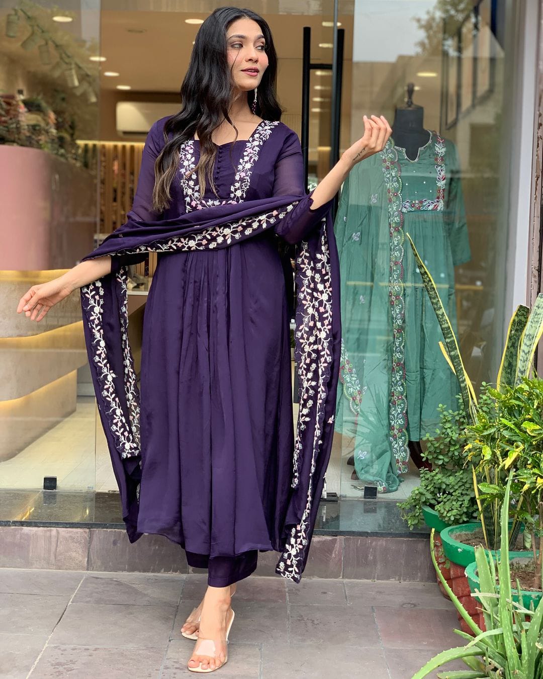 A woman models a deep purple three-piece ethnic suit with white floral embroidery on the neckline, sleeves, and dupatta, standing in front of a glass storefront window.
